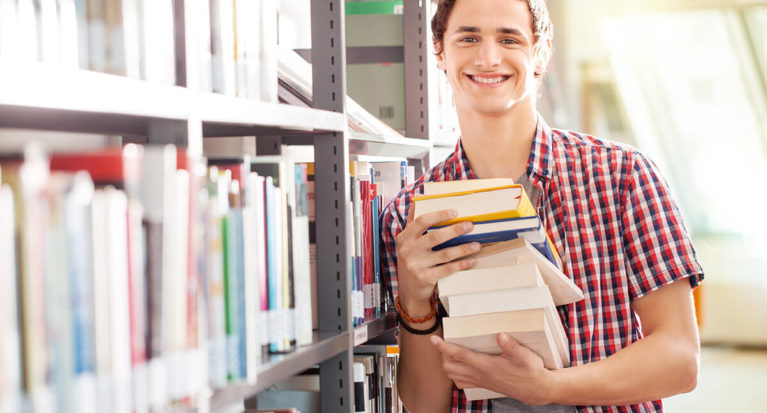 Portrait of a young student holding books standing by bookshelf in library